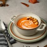 Tasty squash puree soup with fresh cream and toasted bread pieces in bowl on table with black peppercorns on gray background