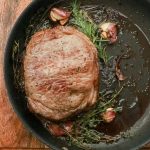 Top view of piece of meat steak in hot pan with rosemary and garlic placed on wooden cutting board in kitchen